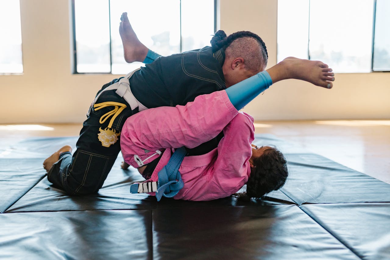 services-03 Two athletes engaged in Brazilian Jiu-Jitsu grappling on mats inside a sunlit gym.
